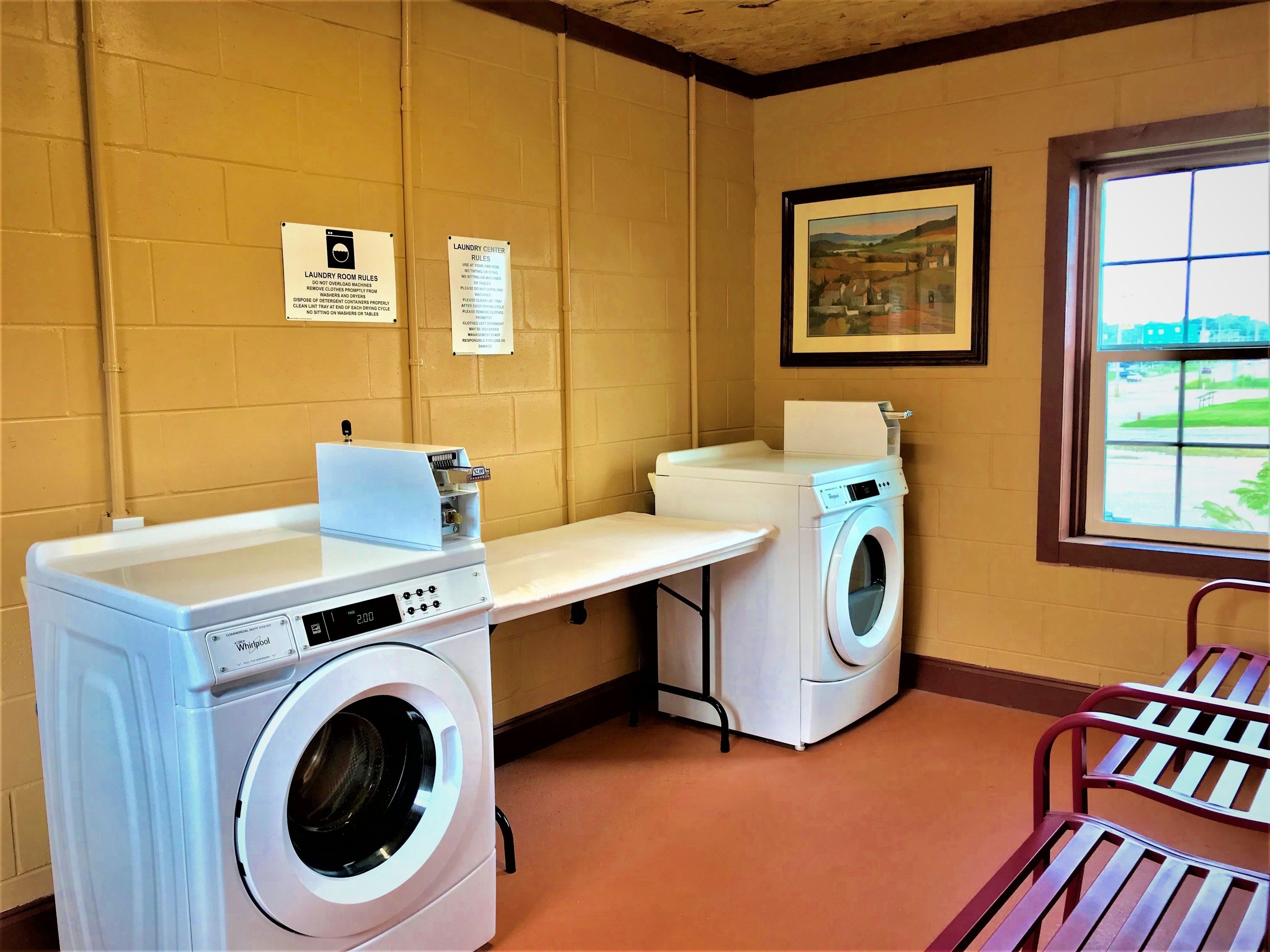 A laundry room with two Whirlpool washers, a drying machine, and a bench. There are signs on the wall with laundry rules.