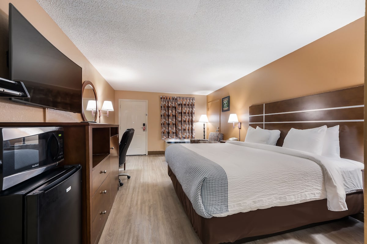 Una habitación de hotel con una cama, un escritorio con computadora, microondas, refrigerador, una silla y televisor.