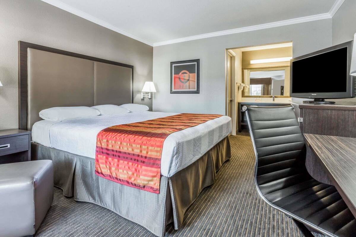 A hotel room with a bed, a desk, a chair, a television, and a bathroom visible through an open door.