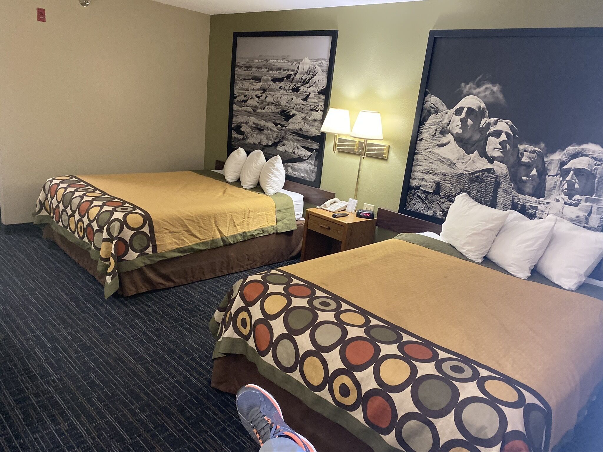 Quarto de hotel com duas camas, uma mesa de cabeceira com abajur, relógio e um quadro na parede.