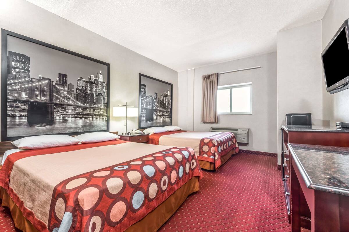 A hotel room with two beds, a desk, a TV, and cityscape artwork on the walls.