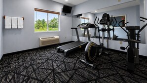 A gym with a treadmill, elliptical, and stationary bike, a large window with blinds, and a ceiling-mounted air conditioner.