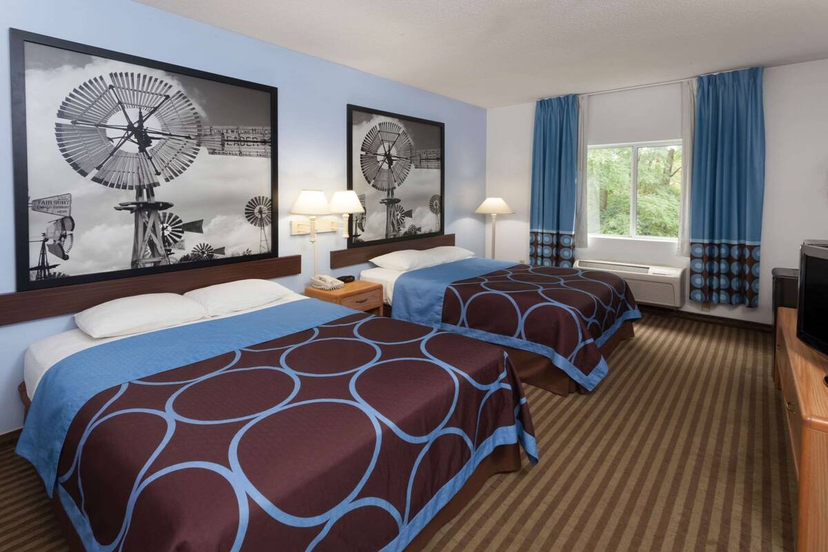 A hotel room with two beds, a TV, a window with curtains, and windmill artwork on the wall.