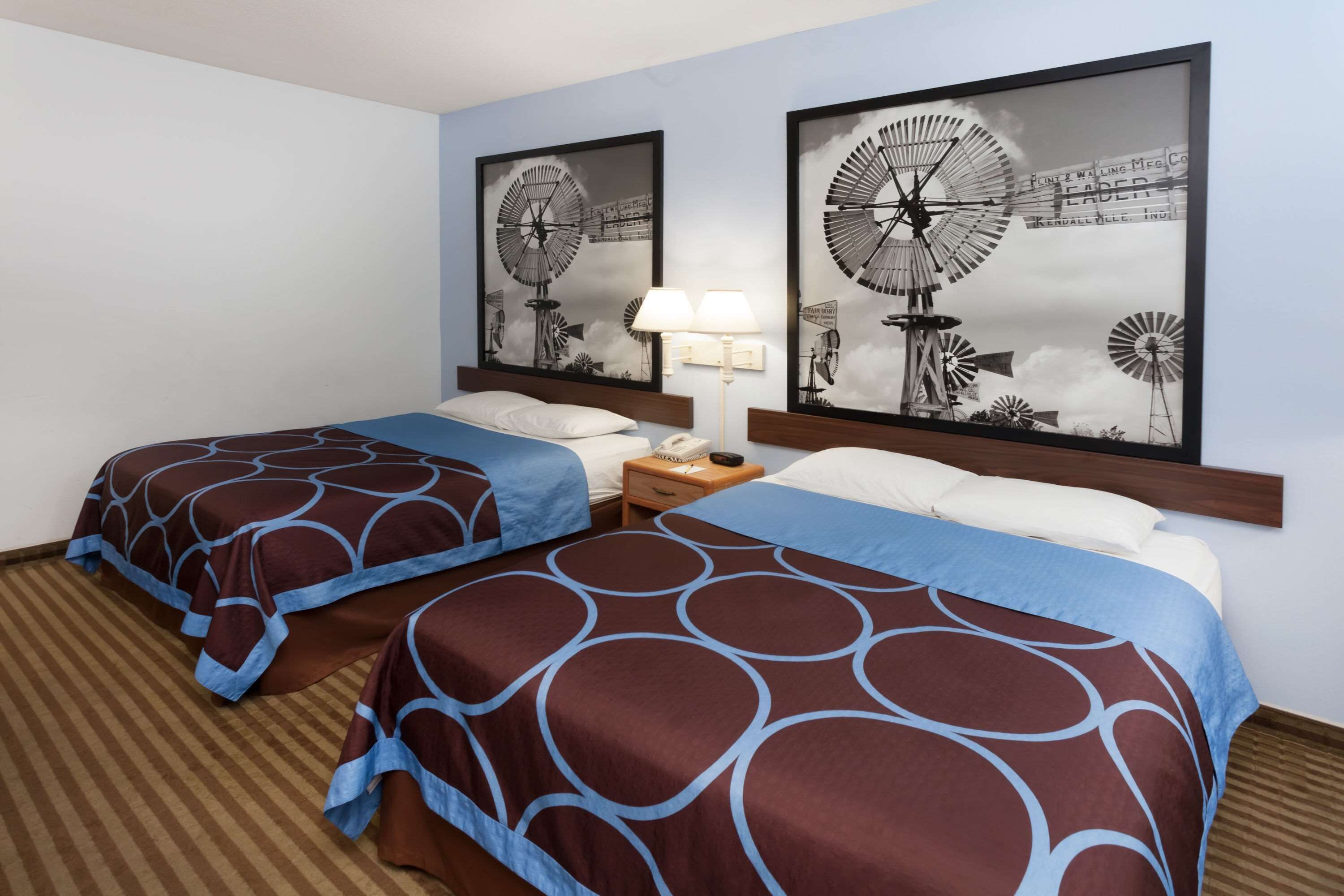 A hotel room with two beds, a nightstand with a lamp, and two framed pictures of windmills on the wall.