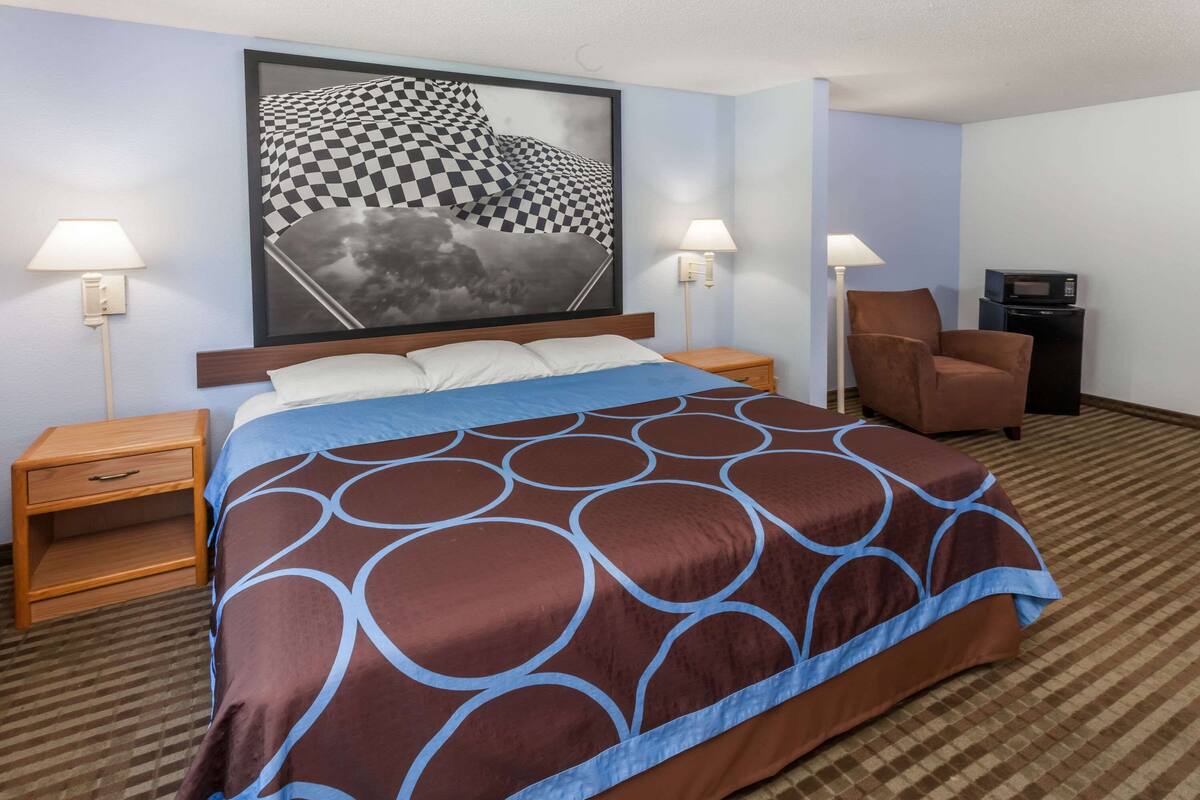 A hotel room with a large bed, two bedside tables, a chair, a desk with a computer, and a wall-mounted TV.