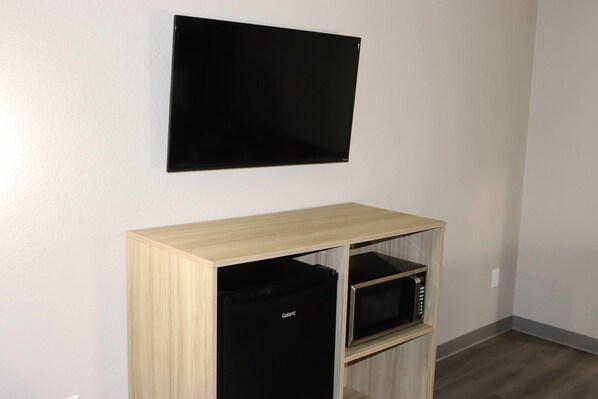 A room with a flat-screen TV mounted on the wall, a wooden cabinet with a microwave inside, and a black appliance below the cabinet.