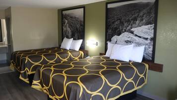 A hotel room with two beds, each with a patterned bedspread, a nightstand, a wall-mounted lamp, and two large framed pictures depicting mountainous landscapes.