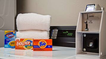 A hotel room with a stack of white towels, a digital clock displaying 2:00, and three boxes of laundry detergent on the table.
