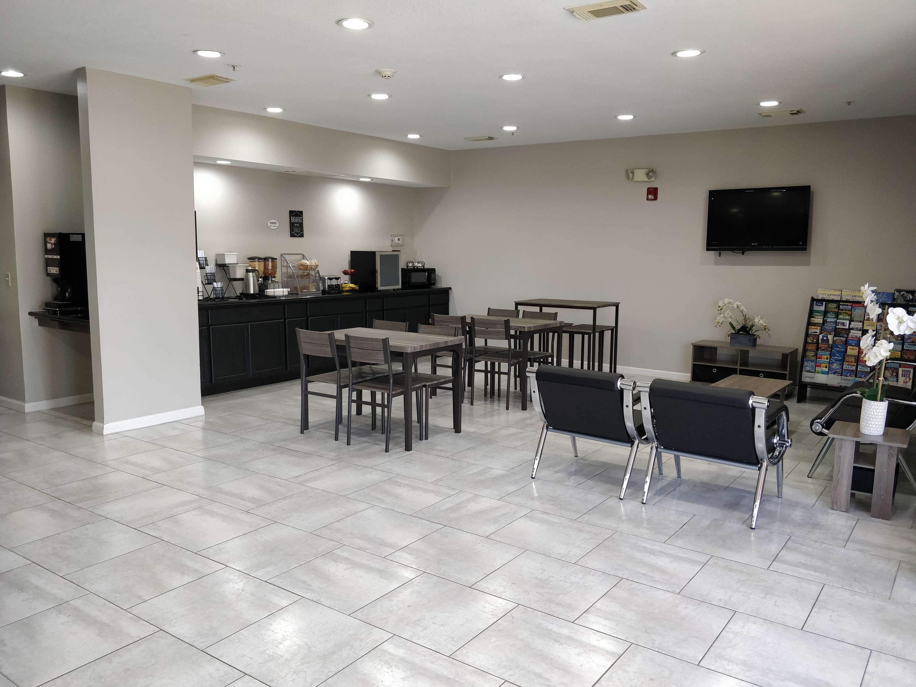 A modern hotel lobby with a dining area, kitchen, and a flat-screen TV on the wall.