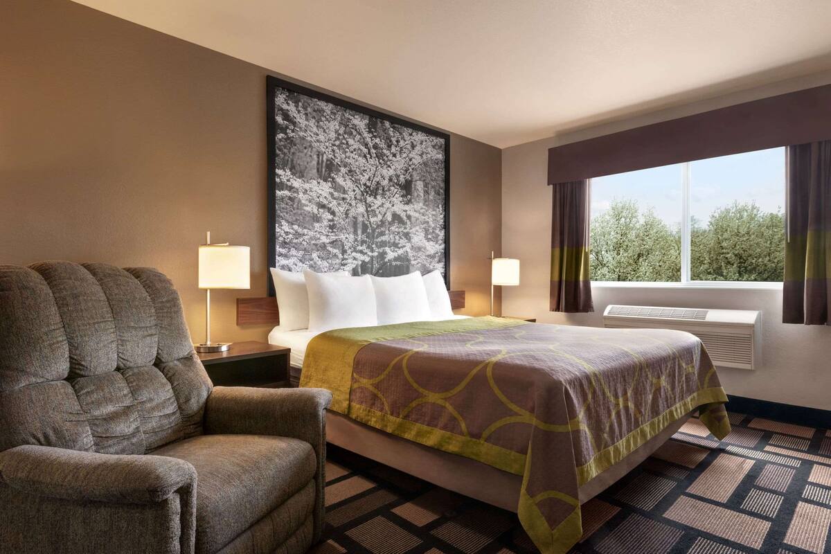 A hotel room with a bed, a chair, a large wall-mounted picture, two bedside lamps, and a window with curtains.