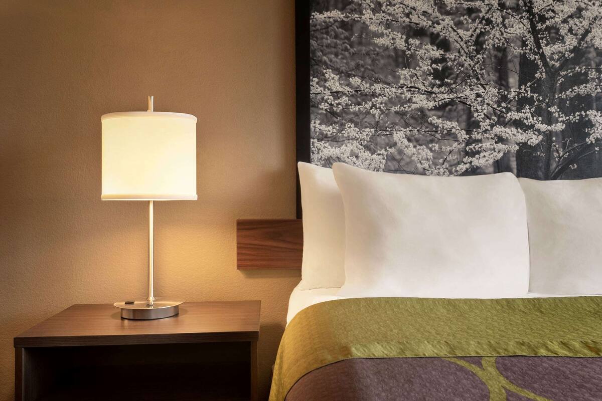 A neatly made bed with white and green bedding, a bedside lamp on a wooden nightstand, and a large framed picture of cherry blossoms on the wall.