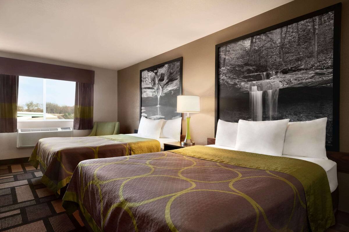 A hotel room with two beds, a large window with curtains, a wall-mounted air conditioner, and two framed pictures of waterfalls.
