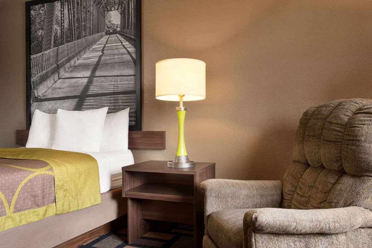 A hotel room with a bed, a nightstand with a lamp, a chair, and a painting on the wall.