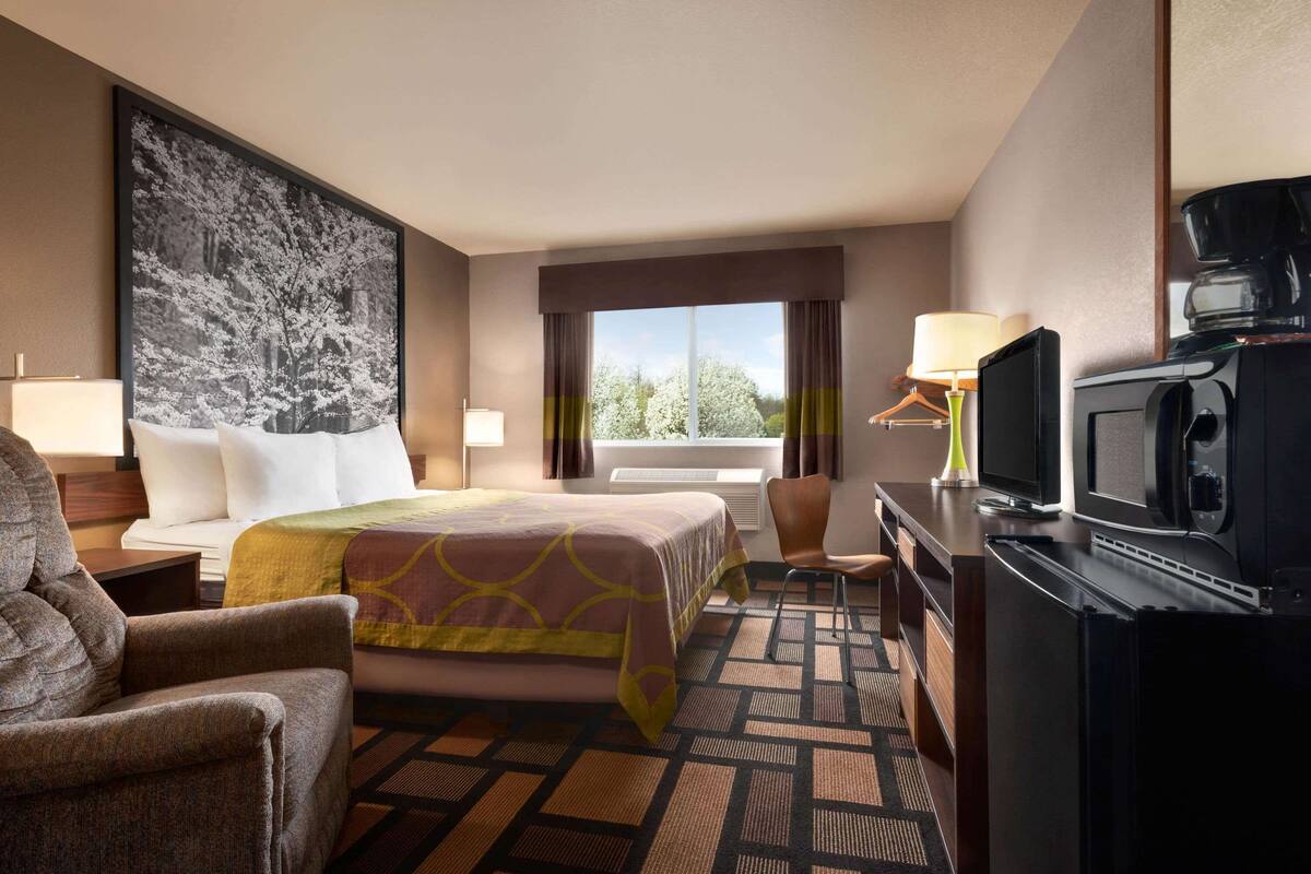A hotel room with a bed, a desk with a television, a chair, a desk lamp, and a large window with curtains.