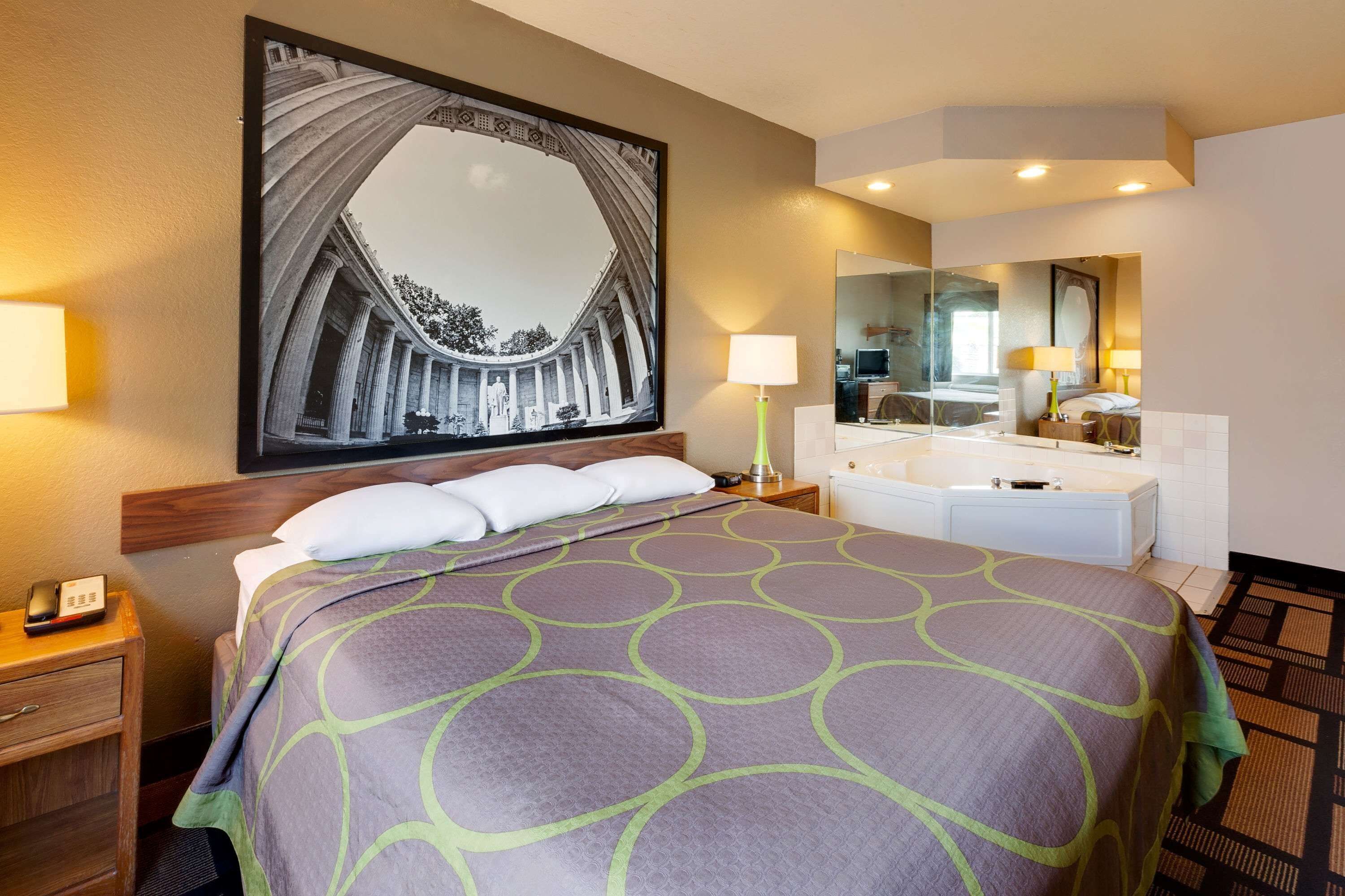 Una habitación de hotel con una cama grande, una mesita de noche con teléfono, un baño con bañera y un espejo amplio.