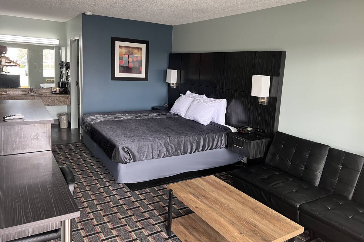 Una habitación de hotel moderna con una cama grande, un sofá, una mesita de madera, una mesita de noche con lámpara, y un baño visible a través de una puerta abierta.