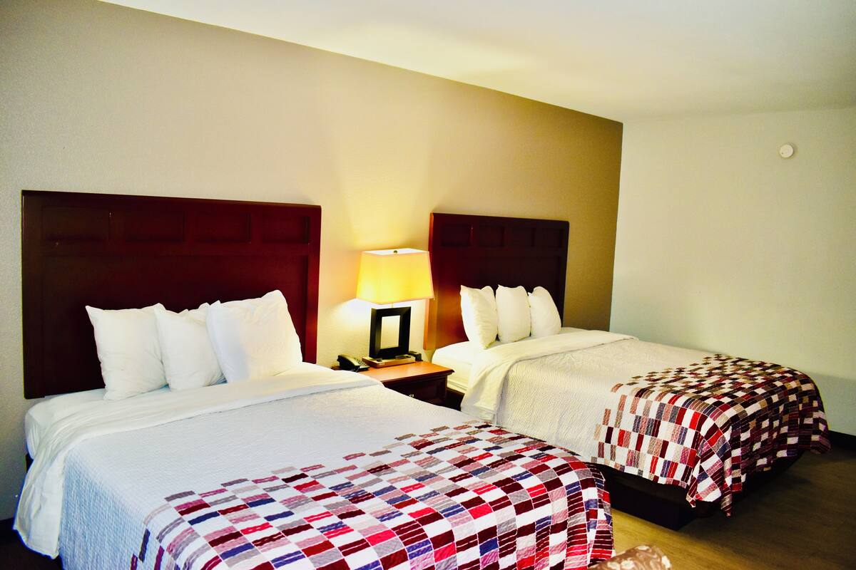 A hotel room with two beds, a nightstand with a lamp, and a plain wall.