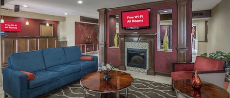 A hotel lobby with a blue sofa, two round tables, a fireplace, and a sign indicating free Wi-Fi in all rooms.