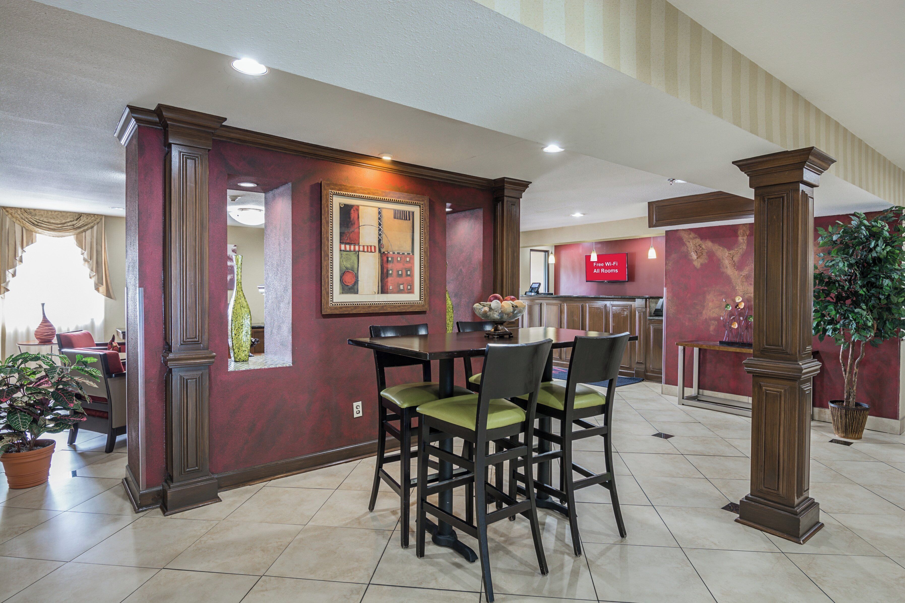 A hotel lobby with a dining area featuring a wooden table and chairs, a reception desk, and a large painting on the wall.