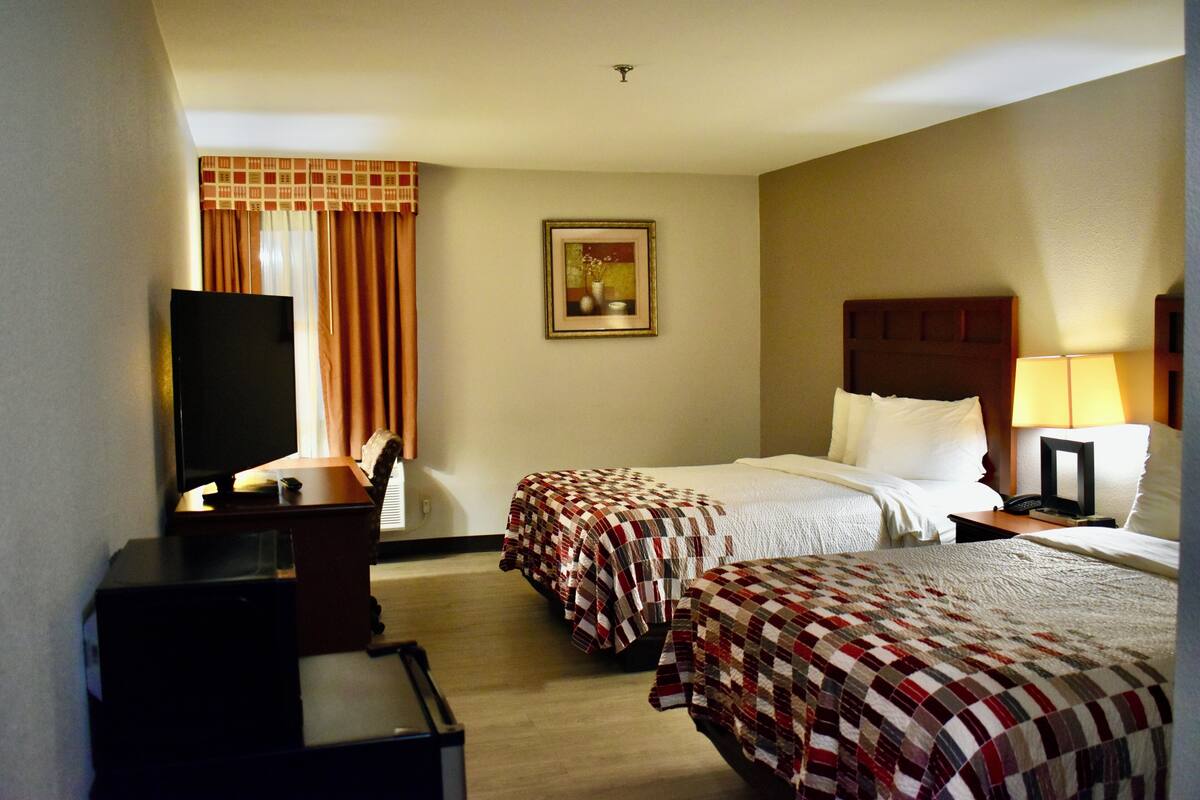 A hotel room with two beds, a desk with a television, a painting on the wall, and a lamp on the nightstand.