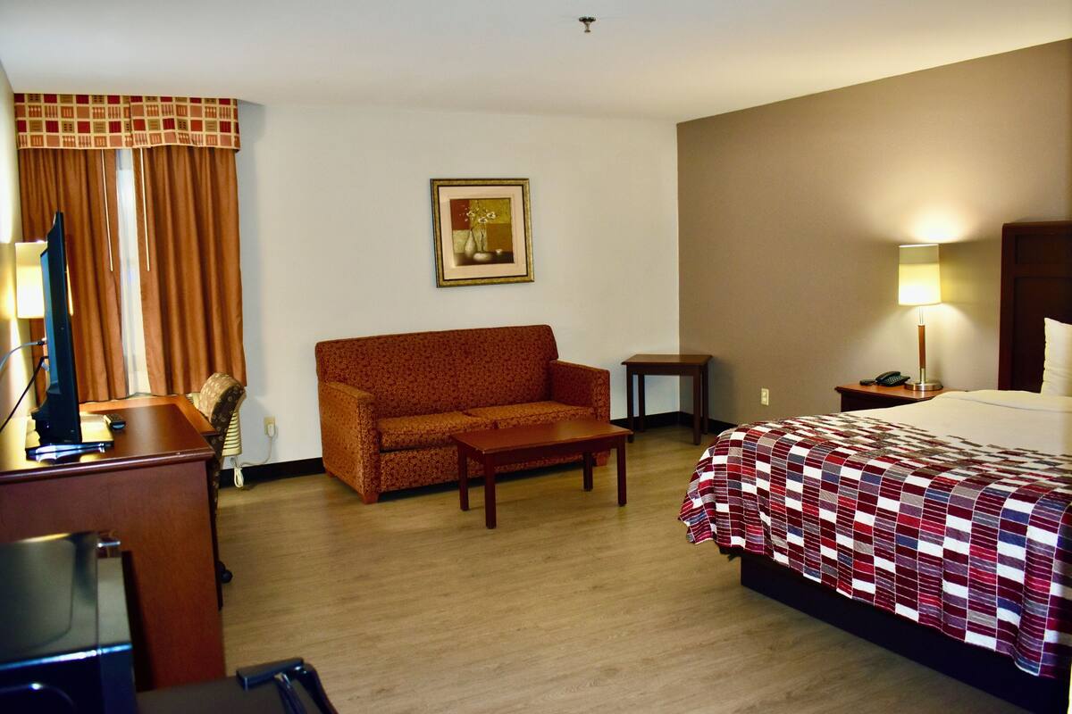 A hotel room with a bed, a sofa, a small table, a desk with a computer, and a lamp.