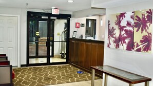 A hotel lobby with a reception desk, a glass door entrance, a patterned carpet, and a wall with a palm tree artwork. - Red Roof Inn & Suites Little Rock (Little Rock)