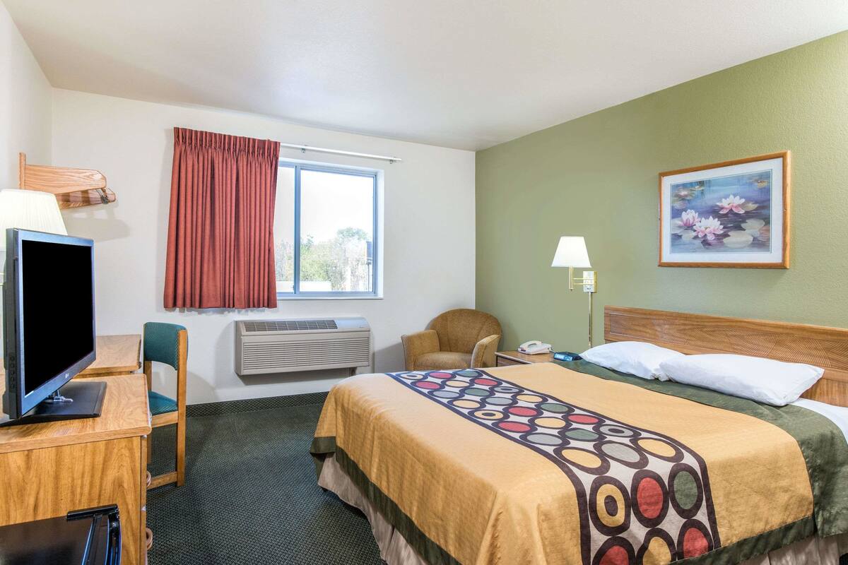 Habitación de hotel con cama, escritorio con computadora, silla, ventana con cortinas, aire acondicionado y un cuadro en la pared.
