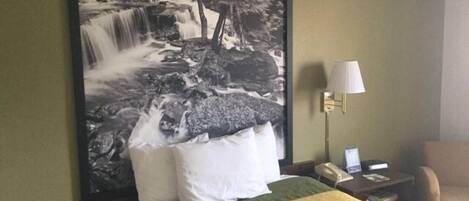 A hotel room with a large waterfall painting on the wall, a bed with green and yellow bedding, a white pillow, a nightstand with a lamp, and a chair.