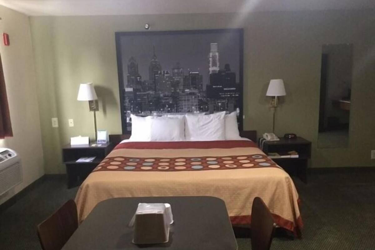 Una habitación de hotel con una cama, dos mesitas de noche, un escritorio con una silla, un cuadro con motivos urbanos y una ventana.