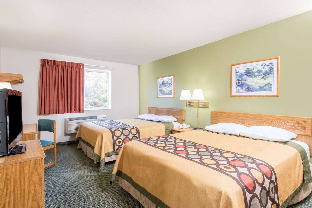 Habitación de hotel con dos camas, un escritorio con una computadora, una silla, una ventana con cortinas y dos cuadros en la pared.