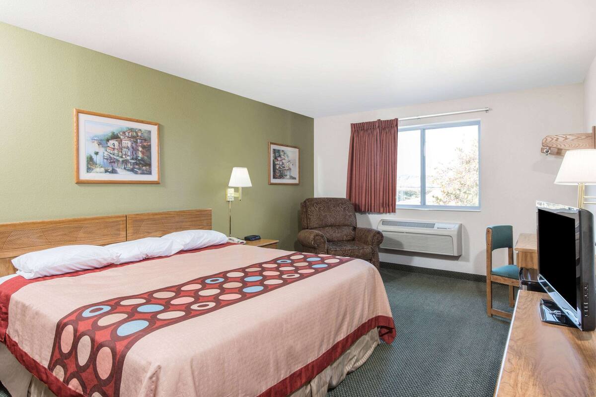 Una habitación de hotel con una cama, una silla, un escritorio con una computadora y dos cuadros en la pared.