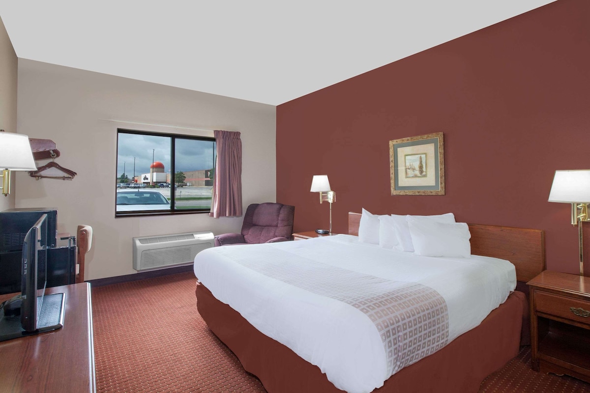 A hotel room with a large bed, a desk with a computer, a chair, a window with a view of a building, and a picture on the wall.