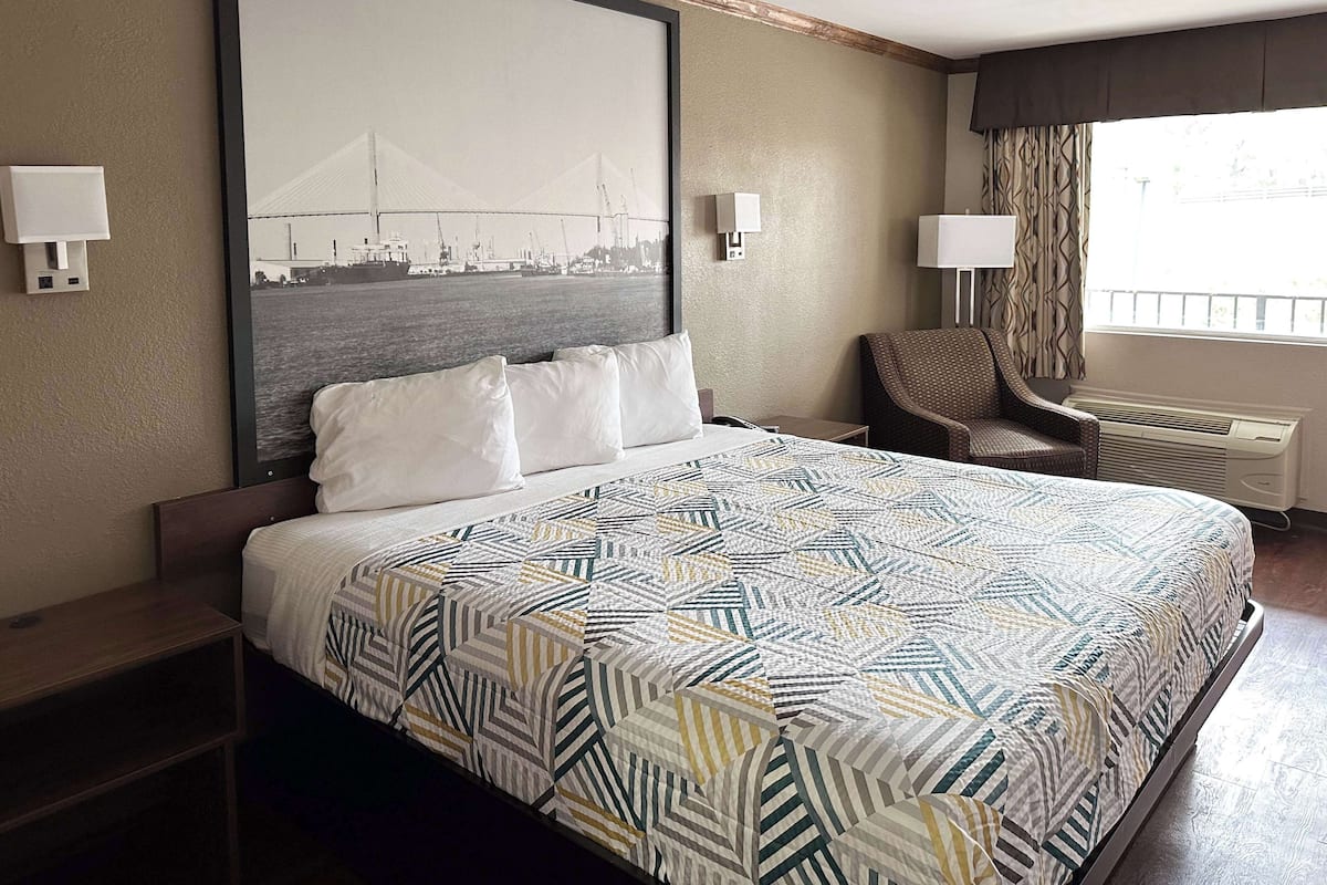 Une chambre d’hôtel avec un grand lit, une chaise, une fenêtre avec des rideaux et un tableau accroché au mur.