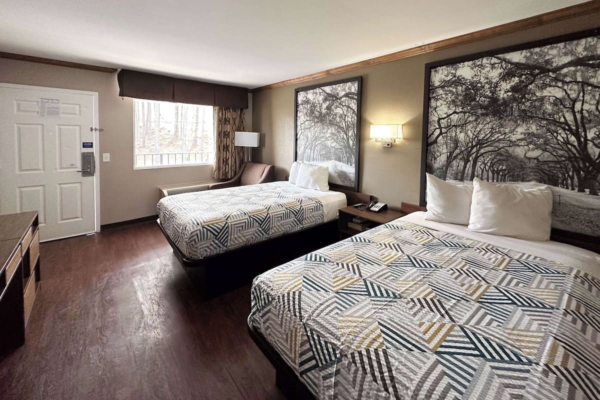 Une chambre d’hôtel avec deux lits, un bureau, une chaise, une fenêtre avec des rideaux, une lampe fixée au mur et un grand tableau sur le thème des arbres.