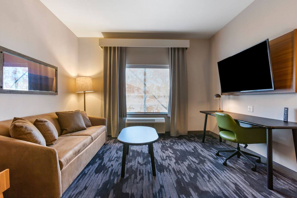 A hotel room with a sofa, a small table, a green chair, a TV, and a window with curtains.