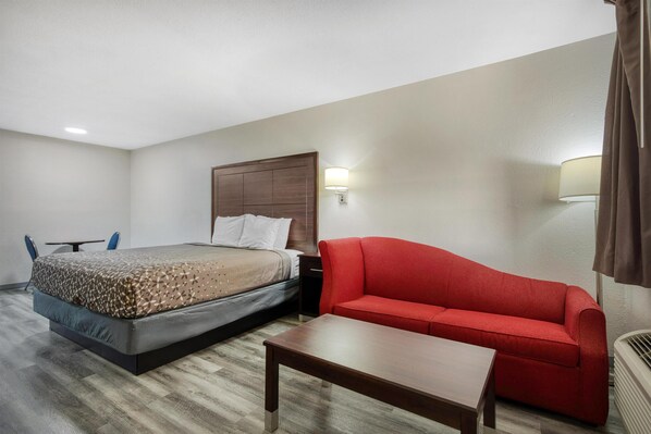 A hotel room with a bed, a red sofa, a wooden table, and a wall-mounted lamp.