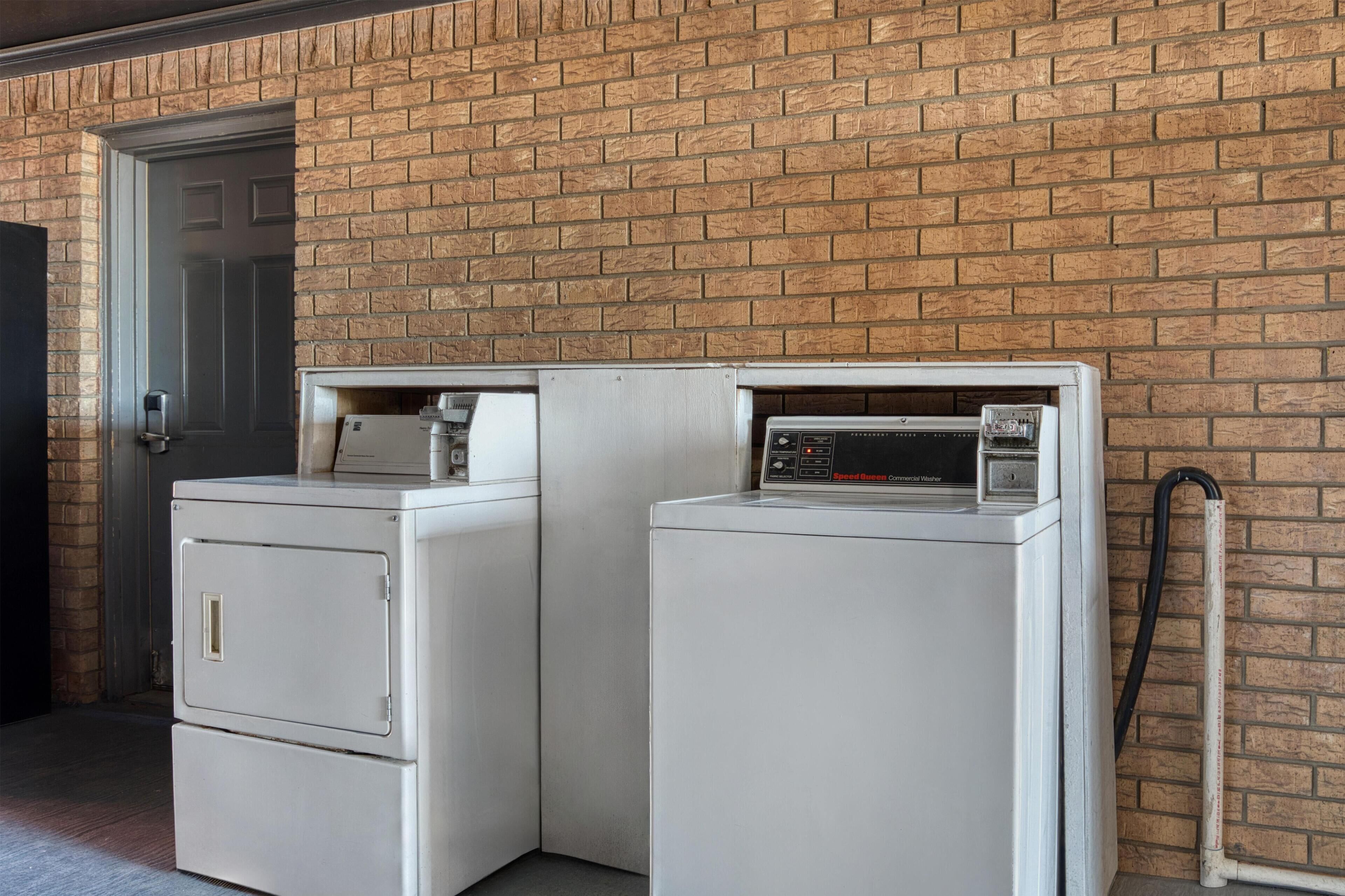 Two white front-loading washing machines against a brick wall, with a door and a hose visible.