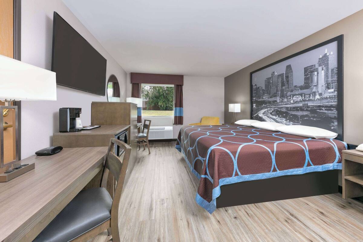 A hotel room with a bed, desk, chair, and a large cityscape painting on the wall.