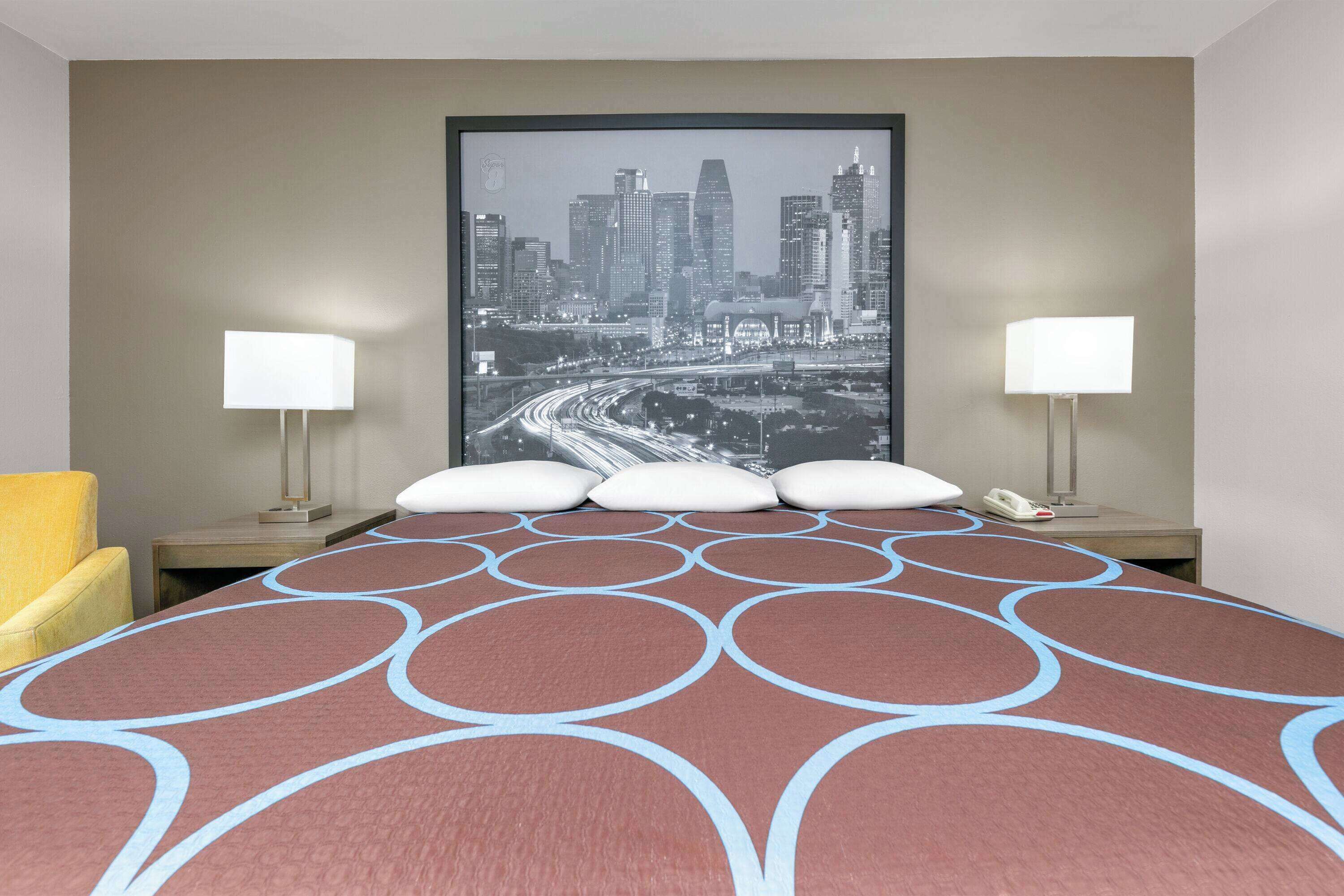 A hotel room with a large bed, two bedside lamps, a cityscape poster on the wall, and a yellow armchair.