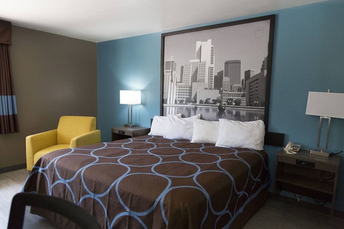 A hotel room with a bed, a yellow armchair, a nightstand with a lamp, and a cityscape painting on the wall.