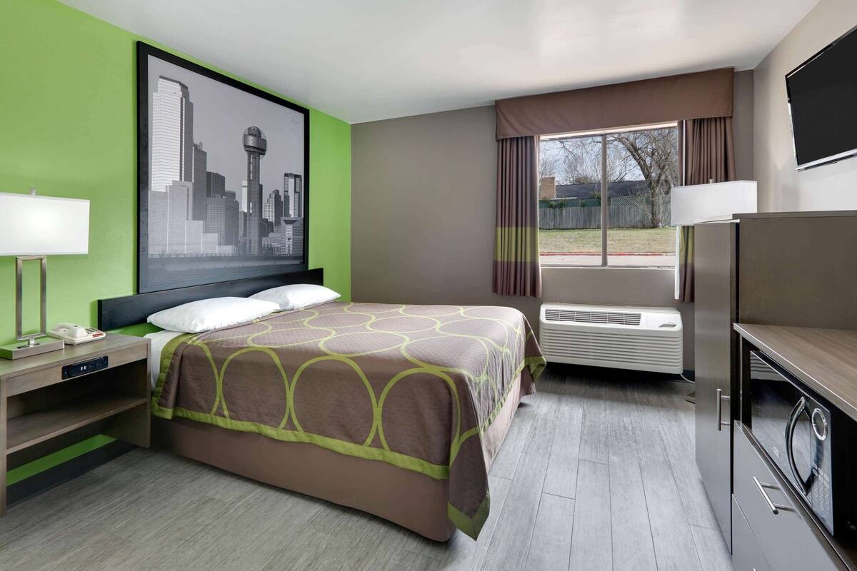 A hotel room with a bed, a flat-screen TV, a desk with a lamp, a large window with curtains, and a cityscape painting on the wall.