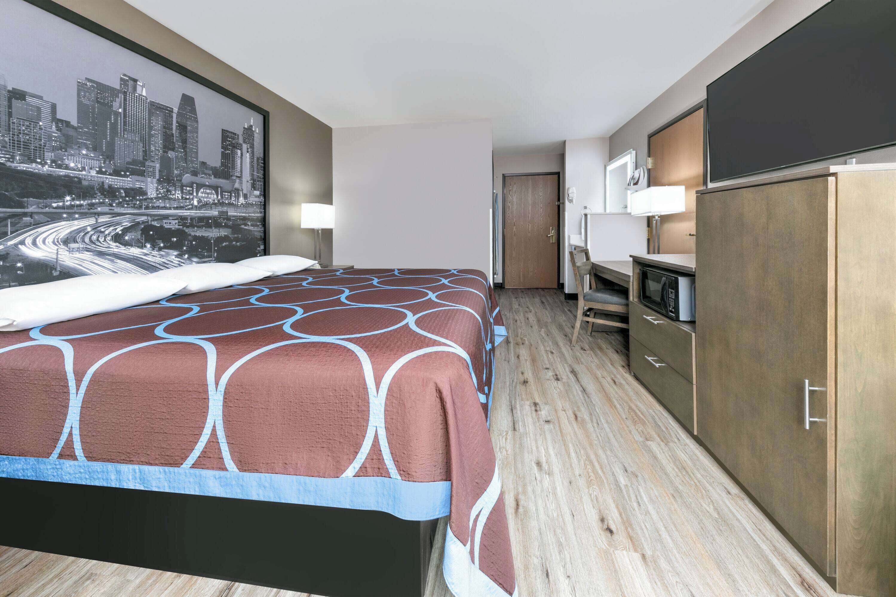 Habitación de hotel con cama, escritorio con computadora, televisión y un cuadro de paisaje urbano en la pared.
