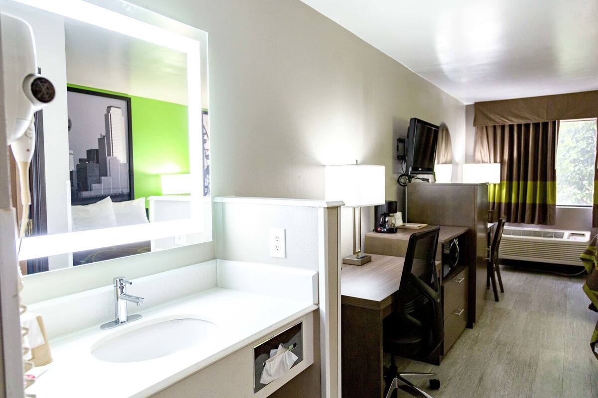 A hotel room with a large mirror, a sink, a desk with a chair, a television, and a window with curtains.