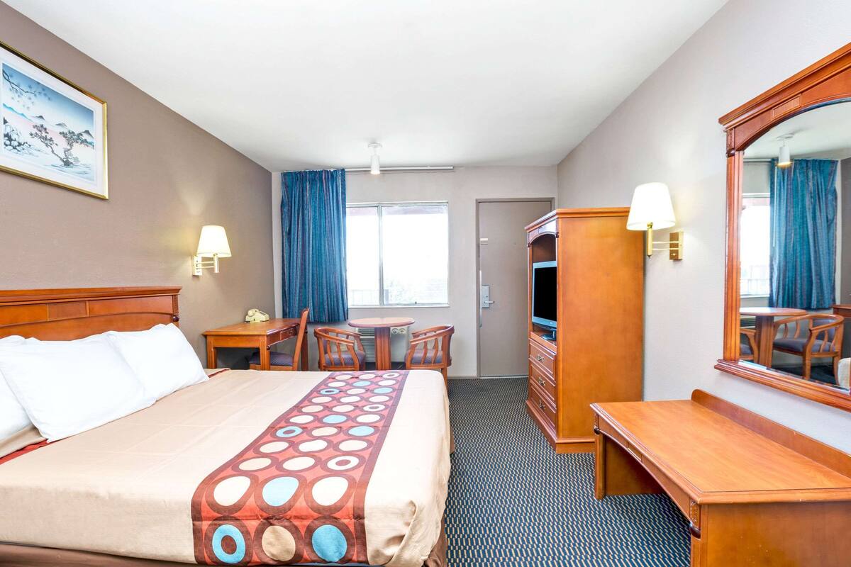 A hotel room with a bed, a desk, a chair, a TV, a lamp, a mirror, and a window with curtains.