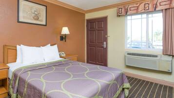 A hotel room with a bed, a nightstand, a lamp, a door, a window with curtains, and an air conditioning unit.