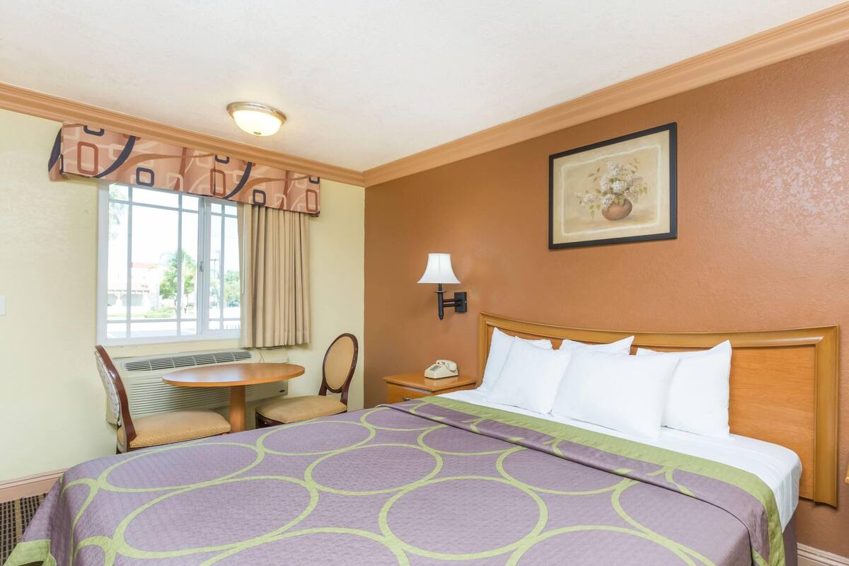 A hotel room with a bed, a desk, a chair, a window with curtains, a lamp, and a framed picture on the wall.