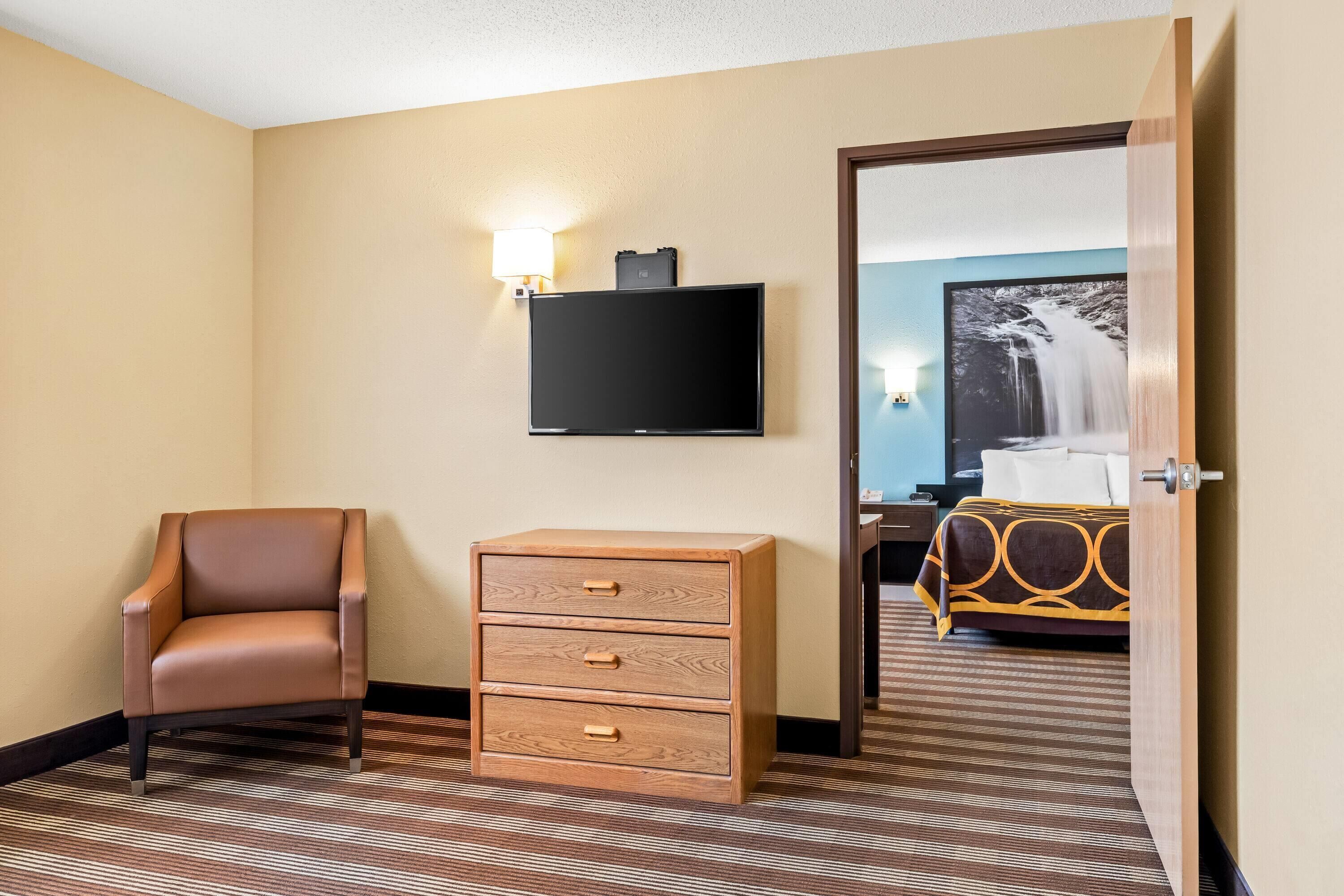 A hotel room with a brown leather chair, a wooden chest of drawers, a flat-screen TV, and a bed with a patterned bedspread.