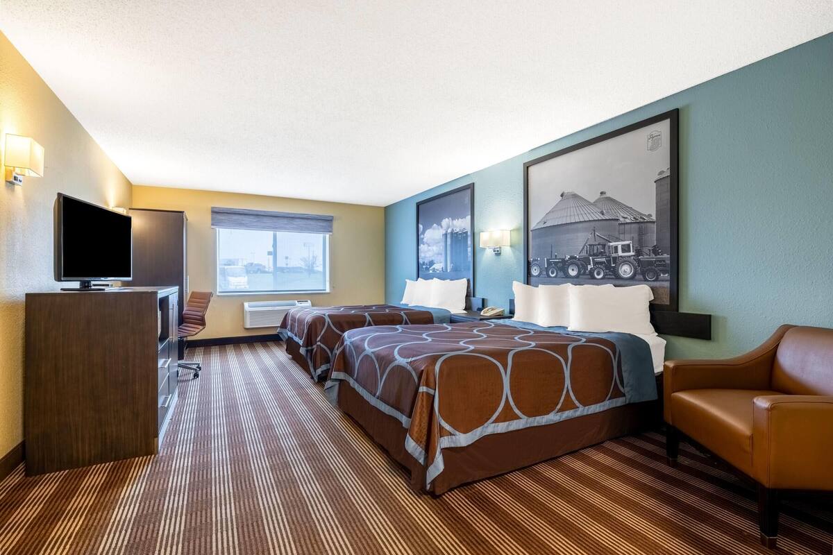 Ein Hotelzimmer mit zwei Betten, einem Schreibtisch mit Fernseher, einem Sessel, einem Fenster mit Stadtblick und einem gerahmten Bild an der Wand.