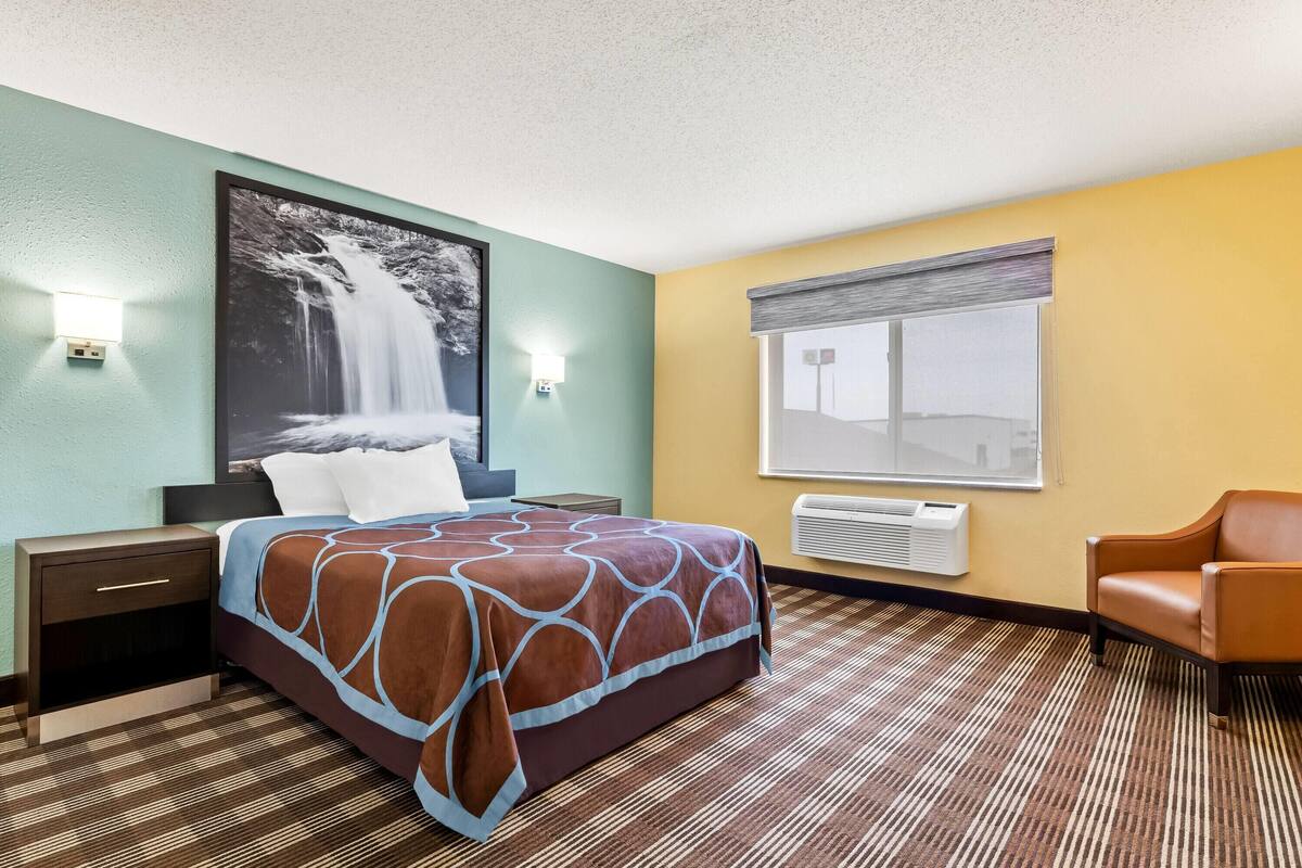 Ein Hotelzimmer mit Bett, Nachttisch, Stuhl, großem Fenster mit Jalousien und einer Wand-Klimaanlage.