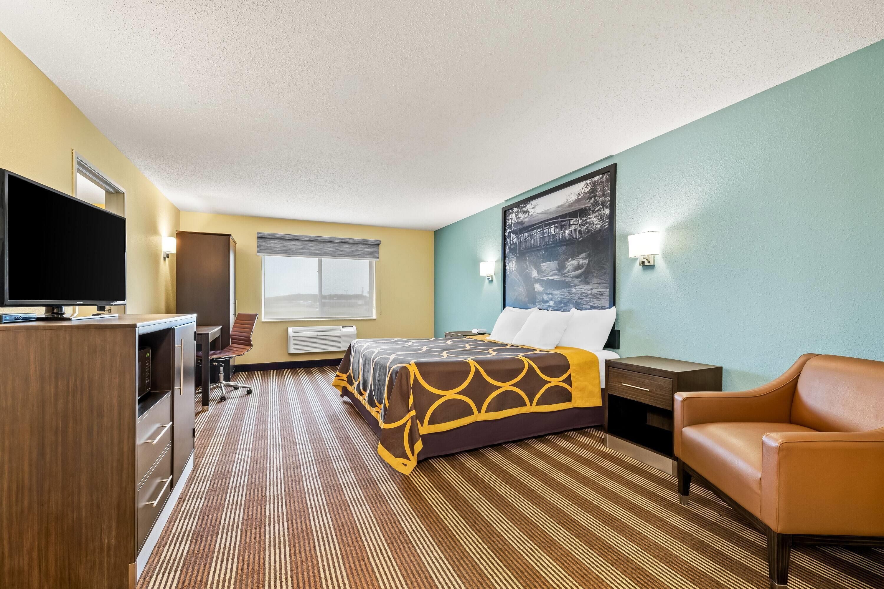 A hotel room with a bed, a desk, a TV, a chair, and a painting on the wall.
